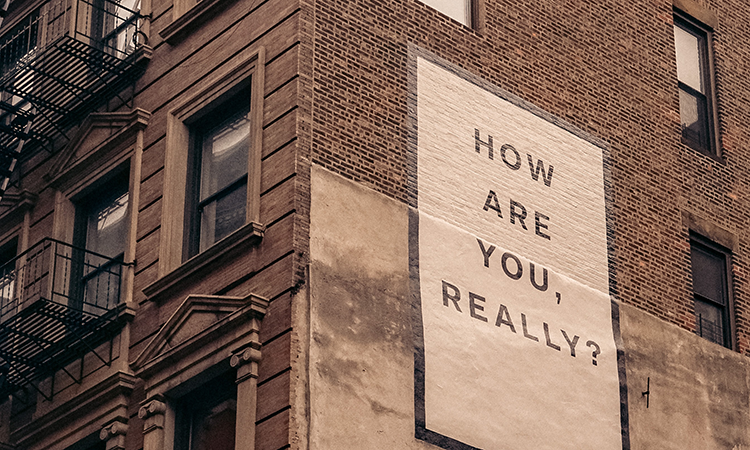 The words "how are you really" printed on a white sign on a brick wall