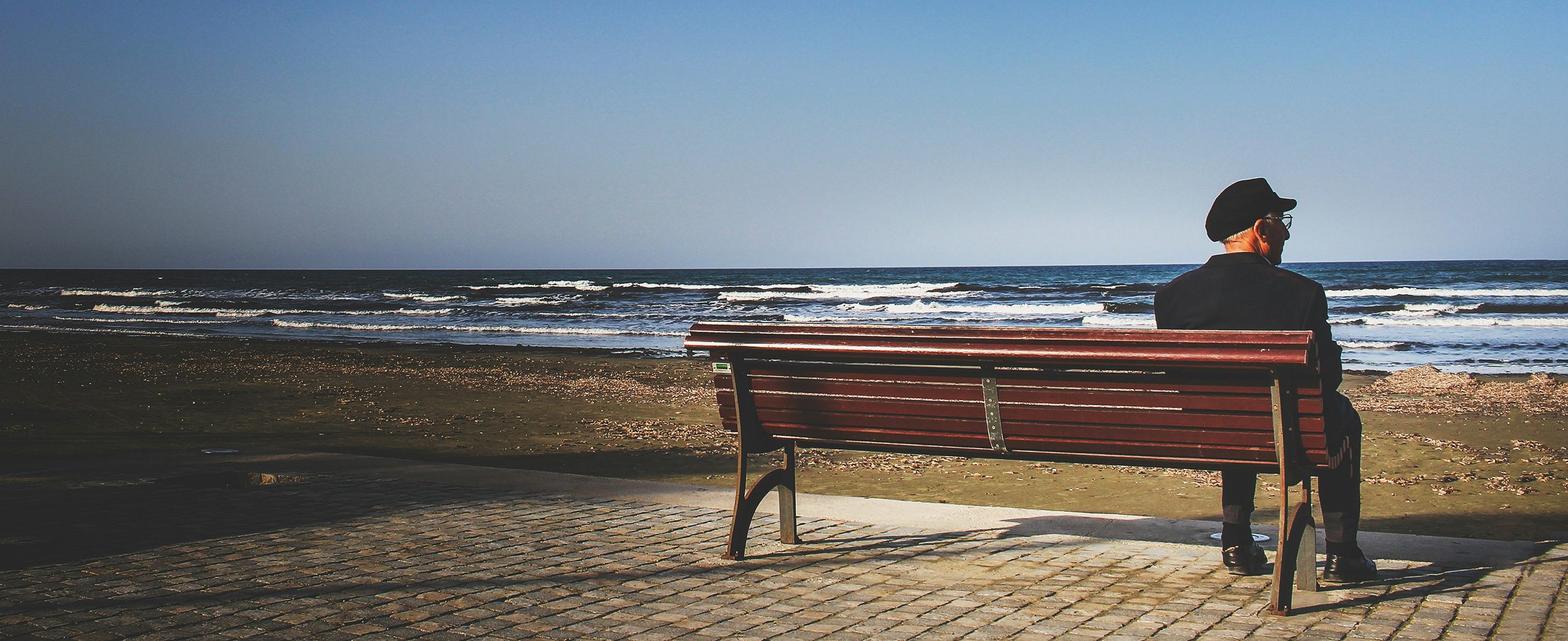 An elderly man sitting alone on a bench in front of the ocean.