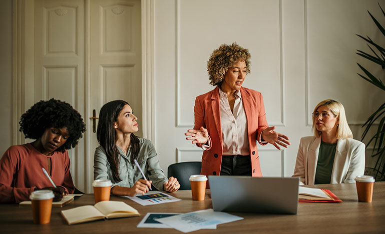 Four professional women working at a large table while one speaks to the group.