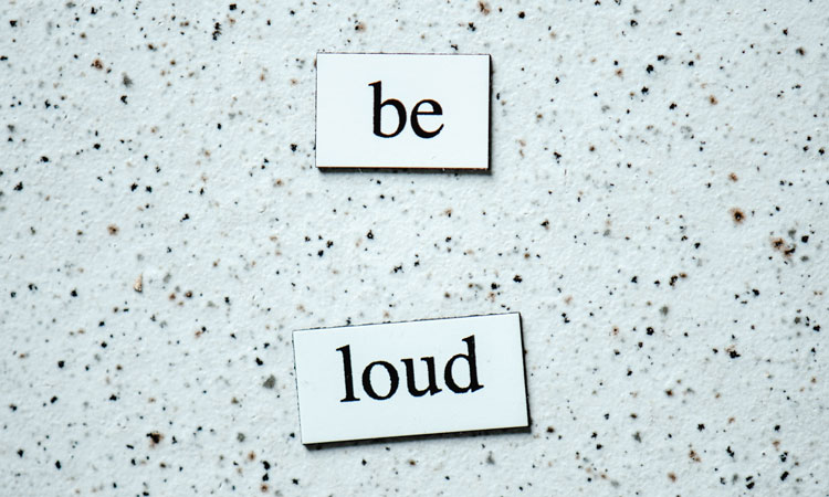 The words "be loud" printed on white magnets on a black speckled white surface