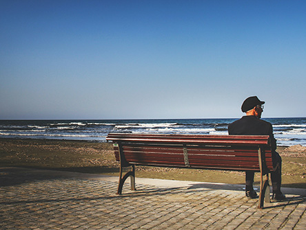 An elderly man sitting alone on a bench in front of the ocean.