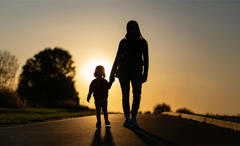 A mother walking and holding her child's hand as they walk towards a setting sun.