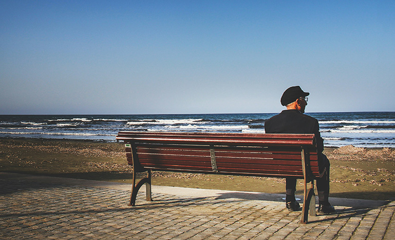 An elderly man sitting alone on a bench in front of the ocean.