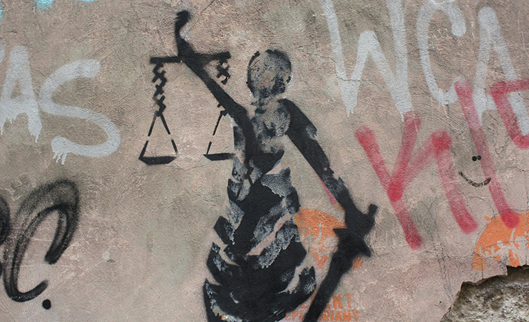 A spray-painted scales of justice surrounded by graffiti on a concrete wall.