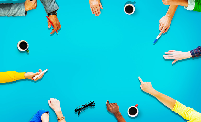 A circle of hands reaching into the middle of an empty table showing collaboration and participation. 